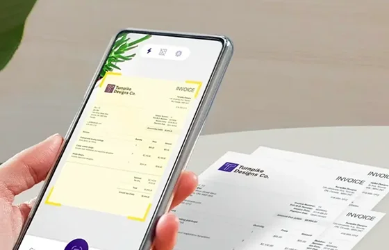 A close-up of a mobile phone scanning a printed invoice from "Turnpike Designs Co." The invoice is digitized on the screen, indicating a feature for document capture or expense tracking.