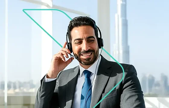 A professional, smiling man wearing a business suit, tie, and a headset with a microphone, representing a dedicated account manager. He is seated in a modern office with a bright window view of a city skyline, including the Burj Khalifa, emphasizing local presence and high-level service.